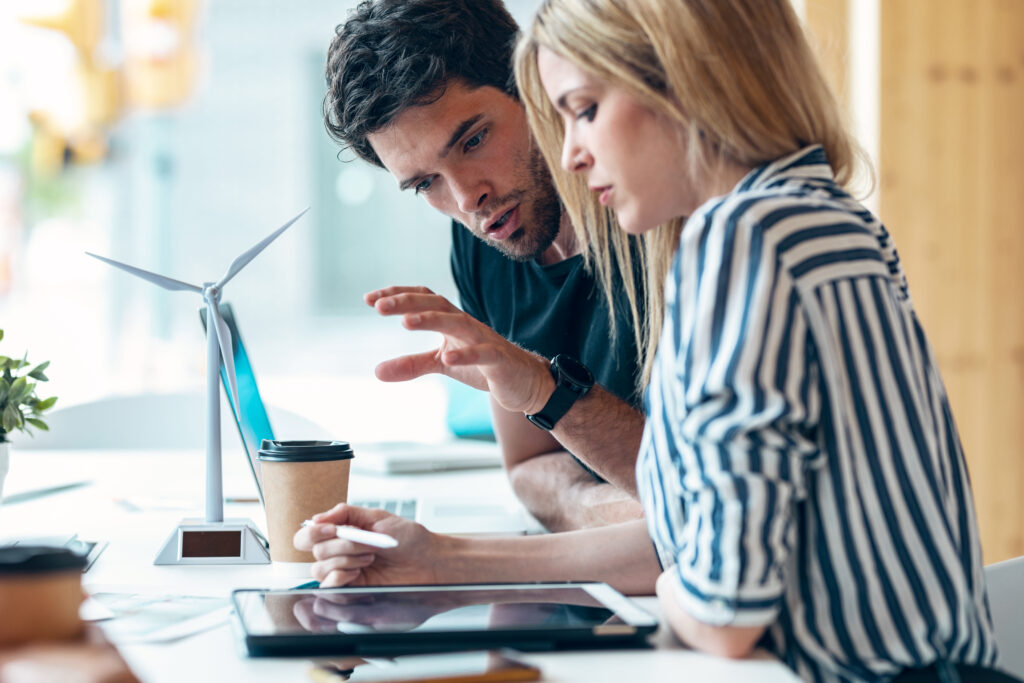 A woman in a shirt and a man sit side by side, bent over a screen analysing data. A miniature wind turbine sits on the desk.