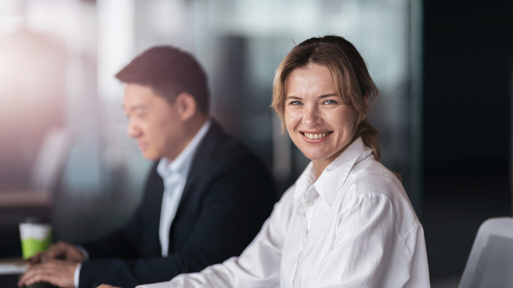 portrait d'une femme blonde et souriante, portant une chemise blanche, elle est tournée vers l'objectif. Dans la profondeur de champ, un homme asiatique en costume sombre.