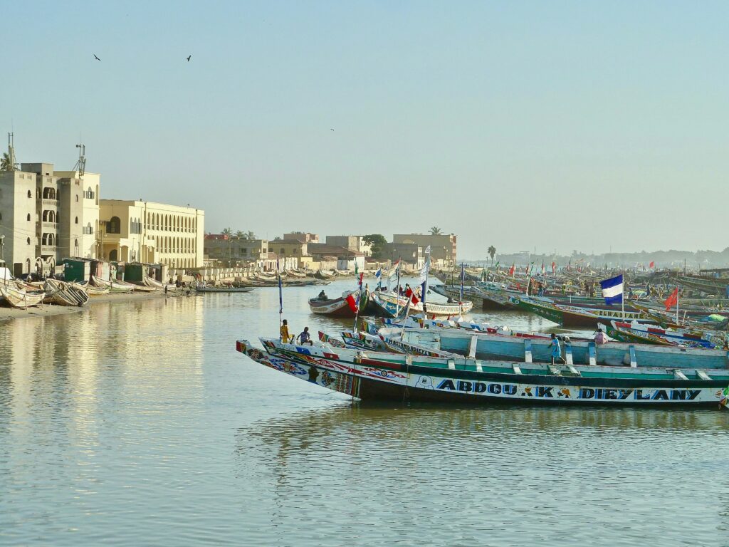 Paysage du Sénégal avec un alignement de barques traditionnelles sur la mer sur la droite et vue sur les bâtiments blancs du front de mer ensoleillé sur la gauche