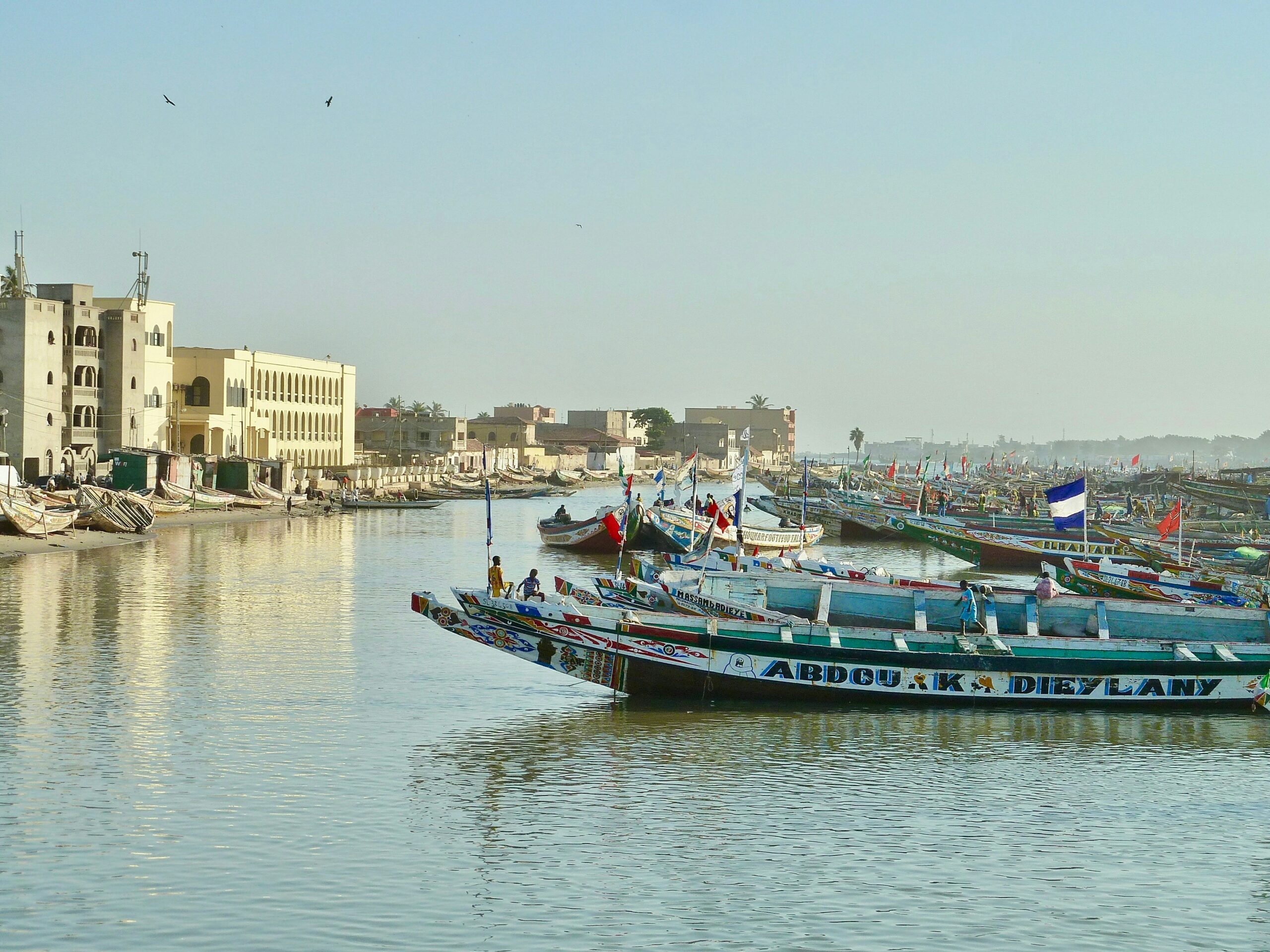 Paysage du Sénégal avec un alignement de barques traditionnelles sur la mer sur la droite et vue sur les bâtiments blancs du front de mer ensoleillé sur la gauche