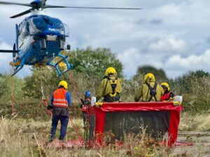 Des techniciens sont dans une nacelle rouge posée dans un champ, un hélicoptère flotte légèrement au-dessus du sol, prêt à emporter la nacelle pour effectuer des travaux sous tension