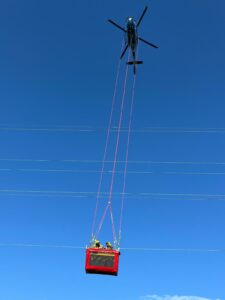 Un hélicoptère RTE transporte une nacelle rouge avec deux techniciens qui effectuent des travaux sous tension sur une ligne électrique